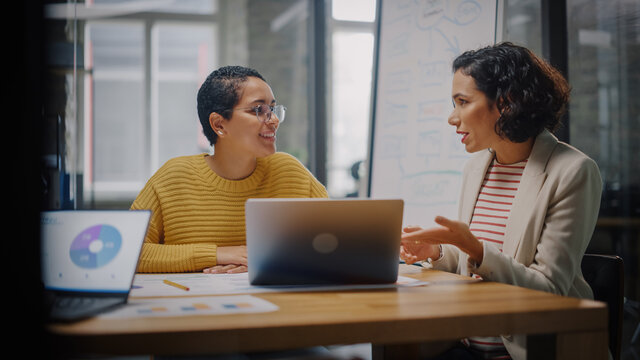 Two Diverse Multiethnic Female Have a Discussion in Meeting Room Behind Glass Walls in an Agency. Creative Director and Project Manager Compare Business Results on Laptop and App Designs in an Office.