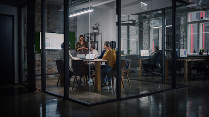 Project Manager Makes a Presentation for a Young Diverse Creative Team in Meeting Room in an Agency. Colleagues Sit Behind Conference Table and Discuss Business Development, Sales Analysis and Design.