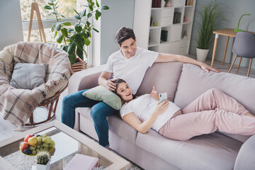 Full size photo of happy young family sit lie sofa hold phone browsing parenthood on weekend in house indoors