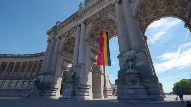 Upwards angle sideways rotating shot of the Cinquantennaire triumphal arc monument in Brussels, Belgium, on warm sunny summer day with blue skies. Belgian flag waving in middle arch.