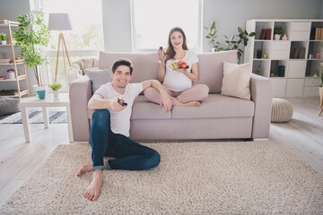 Obraz premium Photo of cheerful positive family waiting baby tummy sitting sofa carpet watching tv eating fruits indoors inside