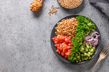Salad of whole grain cereal spelt with fresh summer vegetables, tomato, cucumber, greens on grey table with space for text. View from above.