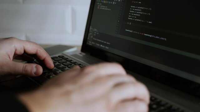 Men's hands press the keys of a laptop keyboard. A man writes a program. The developer is coding. The program code is visible on the screen. Work at home