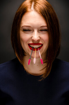 Model with makeup holding a brush for eyelash extension in her teeth, with closed eyes against a dark background.