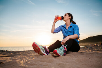 A young pretty woman sits on the sand and drinks a protein shake from a shaker. Bottom view. The...