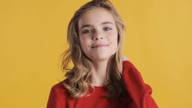 Beautiful cute blond teenager girl with wavy hair dressed in red sweater posing on camera over yellow background