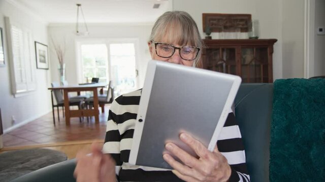 Happy Senior Caucasian woman using tablet computer while sitting in living room. Older white lady using app on e-reader to check news or browse the internet. Slow motion 4k