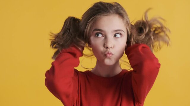 Pretty funny blond teenager girl playing with hair fooling around on camera over yellow background