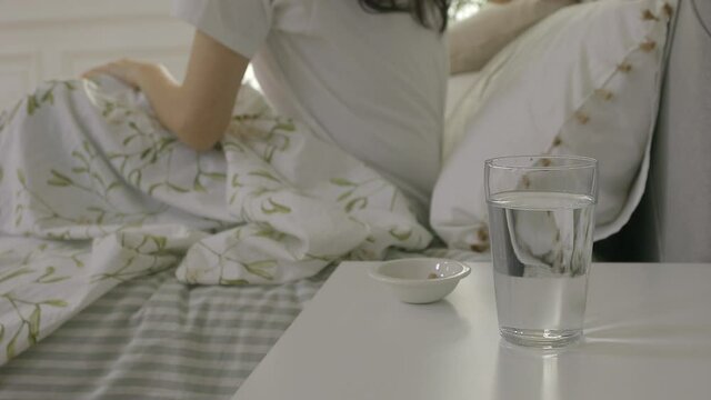 closeup of medicines with water glass on the nightstand with woman sleeping on the bed in background