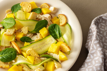 Plate with salad close-up on a brown background, salad of cucumber and chicken breast, mozzarella and croutons with sweet mango pieces and spinach