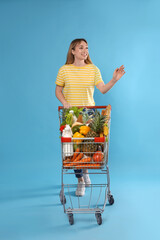 Young woman with shopping cart full of groceries on light blue background