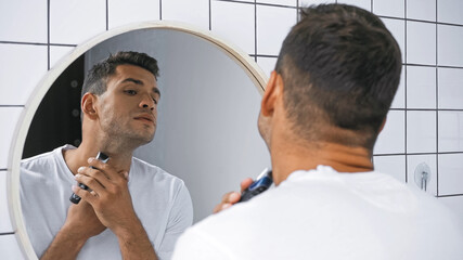 Obraz premium young man shaving neck with electric razor near mirror in bathroom, blurred foreground