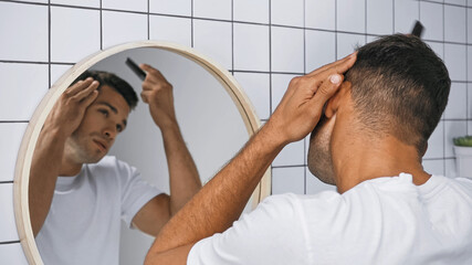 young man brushing hair with comb near mirror in bathroom, blurred background