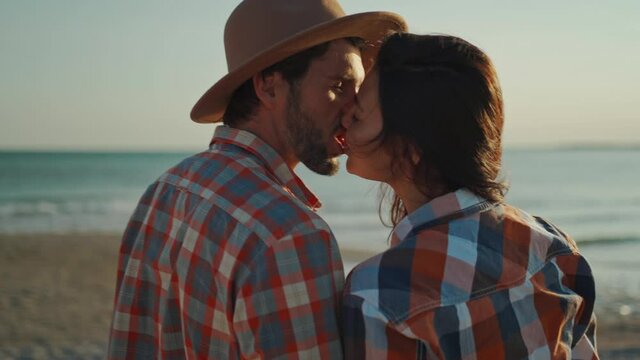 A man wearing a hat and beautiful asian woman kissing on sea beach at sunset. lovely couple travelers in checkered shirts admiring beautiful sea sunset. steadycam shot, camera moving around people