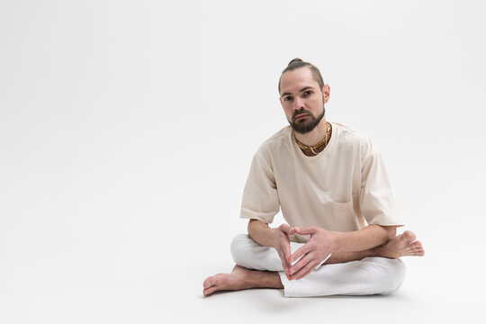 Relaxed young man in white cloath sitting on studio floor isolated on white background