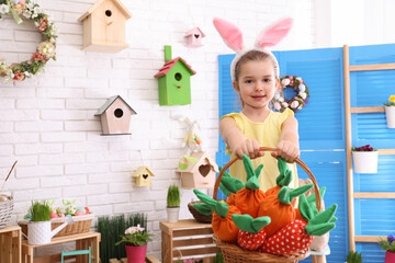 Obraz na płótnie Canvas Adorable little girl with bunny ears and basket full of toy carrots in Easter photo zone