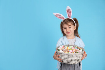 Happy little girl with bunny ears holding wicker basket full of Easter eggs on light blue background. Space for text