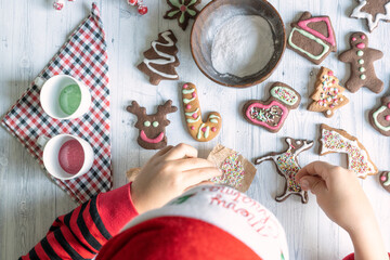 Cute little child with Santa hat decorating the homemade gingerbread cookies at Christmas seen from above