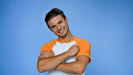 cheerful man smiling at camera while standing with crossed arms isolated on blue