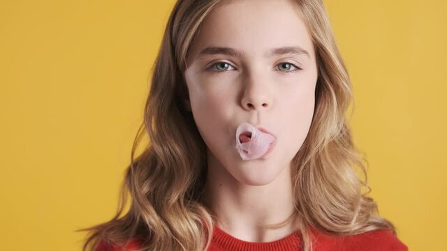 Portrait of pretty blond teenage girl blowing bubble from gum over yellow background. Teen girl blowing candy bubble