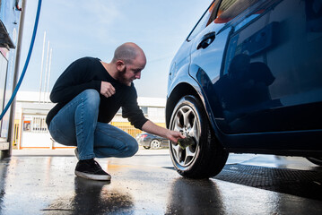 Lavado de coches. Hombre joven limpiando la suciedad de la llanta de una rueda de su coche con una...