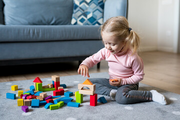 Cute Caucasian baby girl playing with colorful wooden blocks on the floor of the living room at home. Activities with children inside, early childhood development