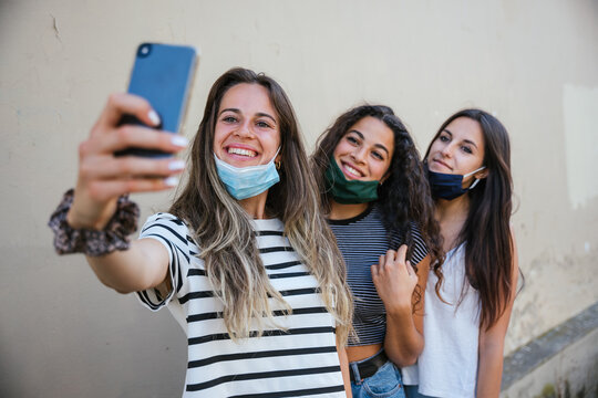 Smartphone selfie portrait three young beautiful women in city at weekend during global coronavirus Covid-19 pandemic with face masks pulled down under noses Millennials have fun together taking photo