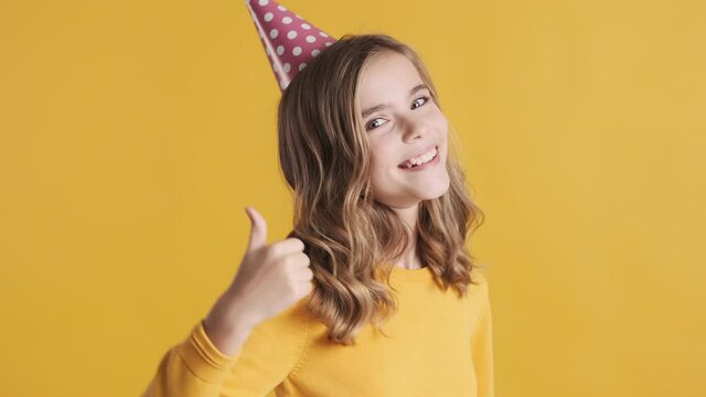 Pretty cheerful blond teenage girl in party hat looking happy keeping thumb up on camera over yellow background
