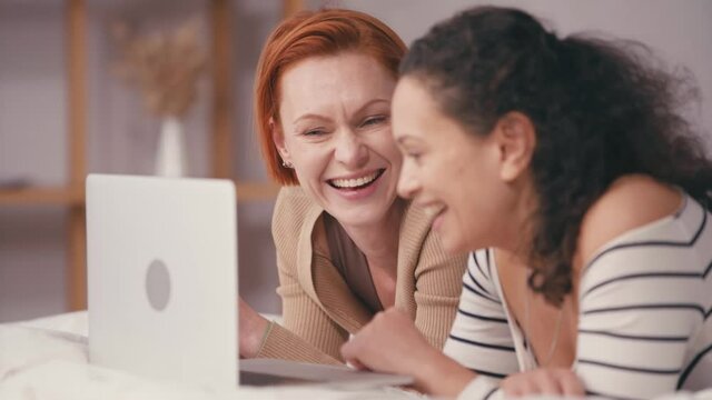 happy redhead woman watching movie on laptop with blurred hispanic girlfriend