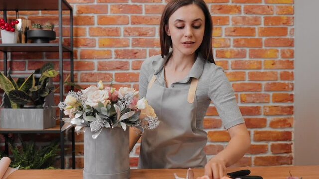 Young woman florist in apron at workplace in flower shop. Brunette girl making bunch of flowers in flowers box. Smiling female florist professional makes bouquet. Composition in hatbox. Slow motion.