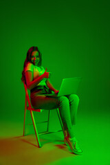 Caucasian woman's portrait isolated on green studio background in multicolored neon light. Typing on laptop, thumb up. Concept of human emotions, facial expression, sales, ad, fashion. Copyspace.