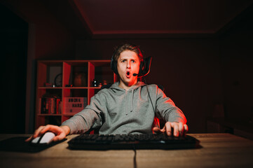 Portrait of a surprised young male gamer in casual clothes and headset sitting in a room with red light at night and looking at the camera with a shocked face. © bodnarphoto