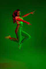 Fototapeta na wymiar Jumps high. Caucasian woman's portrait on green studio background in multicolored neon light. Beautiful female model. Concept of human emotions, facial expression, sales, ad, fashion. Copyspace.