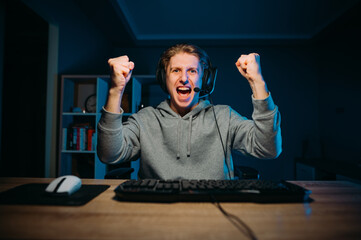 Portrait of a joyful gamer guy sitting at a table at home and rejoices in winning the game with his hands up and looks at the camera and shouts. Gaming concept - obrazy, fototapety, plakaty