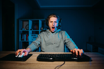 Surprised young male gamer in headset playing video games on computer at home and looking at camera with shocked face. Emotional gaming