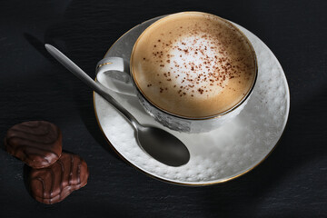 Morning coffee. White cup of cappuccino on a black table. Italian cappuccino. Chocolate chip cookies. Black textured table. Morning dessert in the sun. Italian breakfast.