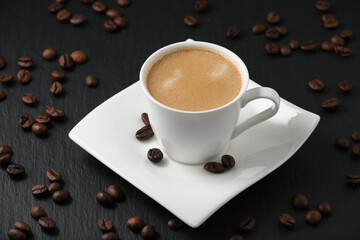 Coffee beans on a black table. White cup of coffee on a black background. Close-up side view, macro photography.