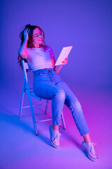 Caucasian woman's portrait isolated on blue studio background in multicolored neon light. Beautiful model with tablet. Concept of human emotions, facial expression, sales, ad, fashion. Copyspace.