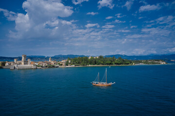 Obraz premium The Old Sailing Ship is sailing near Sirmione Castle. Rocca Scaligera Castle in Sirmione Lake Garda Italy. Aerial view. Clouds in the blue sky.