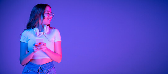 Caucasian woman's portrait on blue background in multicolored neon light. Beautiful model with headphones, phone. Concept of human emotions, facial expression, sales, ad, fashion. Copyspace. Flyer