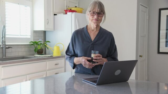 Dolly shot of older white woman wearing glasses with laptop computer on kitchen counter holding iced coffee. Medium shot of Senior Caucasian lady working on notebook pc. Slow motion 4k