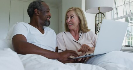 Diverse senior couple sitting in bed using laptop talking and laughing - Powered by Adobe