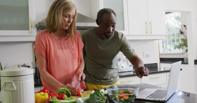 Diverse senior couple preparing food in kitchen following recipe on laptop