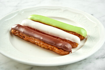 Set of assorted French custard eclairs with custard and assorted toppings served on a white plate on a marble background. Restaurant baking.