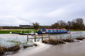 Canal boat stuck trapped in flooded high river overflowing lock with water flowing at speed into...