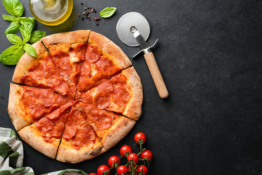 Hot Pepperoni Pizza On Black Concrete Background With Pizza Cutter, Basil Leaf And Cherry Tomatoes On Side. Top View. Delivious Pizza, Copy Space For Text Menu Or Design
