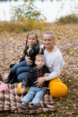 Fototapeta premium A young happy mother with two small children is having picnic in autumn park with colorful leaves.