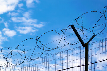 Silhouette of a metal fence with barbed wire from above against the blue sky.