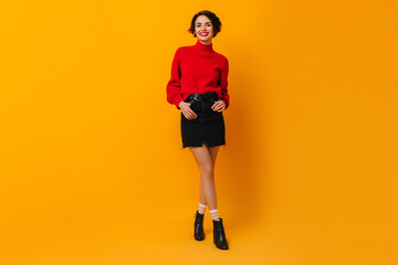 Fototapeta na wymiar Laughing woman in short skirt standing with hands in pockets. Studio shot of cheerful caucasian girl in red sweater.