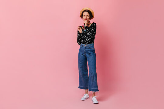 Lovely short-haired woman in straw hat standing on pink background. Full length view of joyful girl in denim pants.